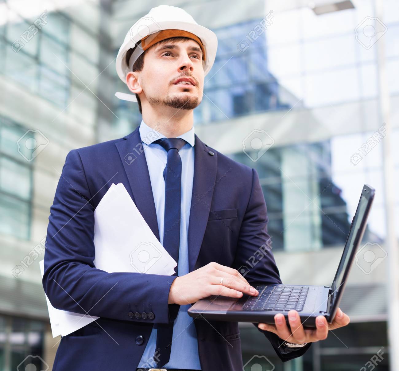 Smiling cheerful  man with documents in suit and helmet working at the laptop